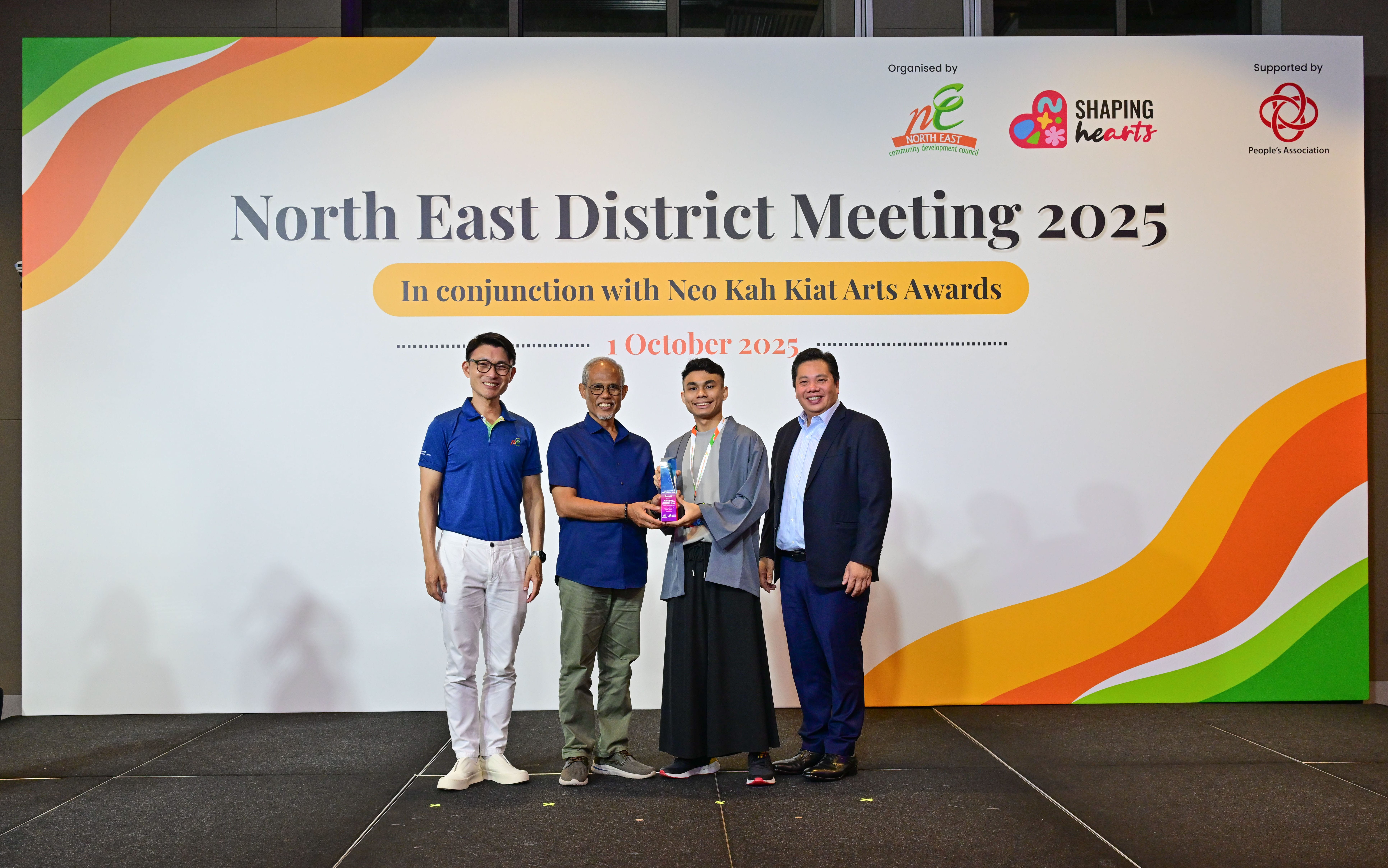 Award recipient standing on stage with the presenter, holding a acrylic plaque and posing for a group photo during the award ceremony, with a backdrop reading ‘North East District Meeting 2025 In conjunction with Neo Kah Kiat Arts Awards'